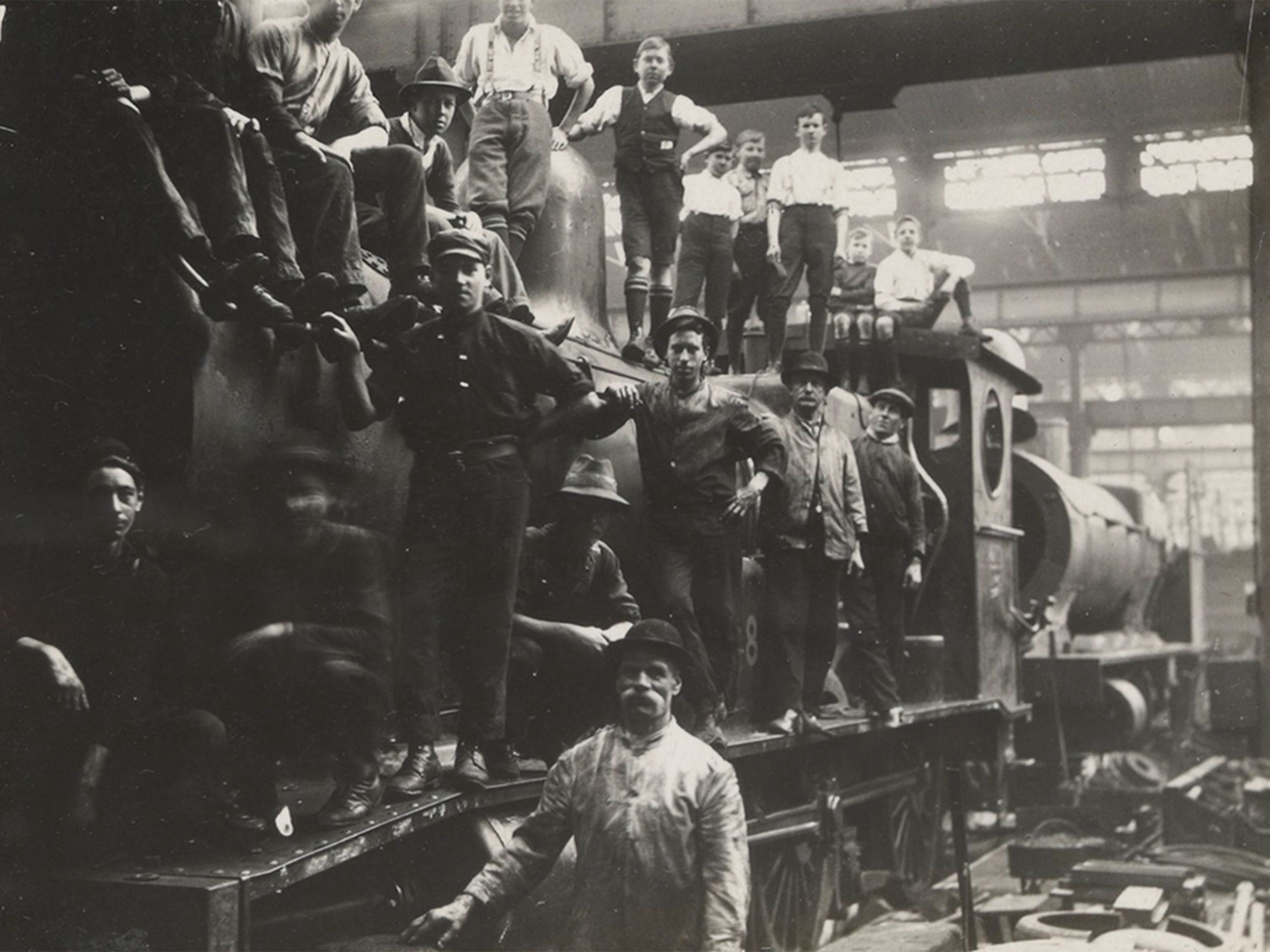 Young men and boys hang off a train posing for the photograph in a large workshop.